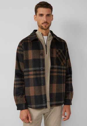 Man wearing a black and brown plaid button-up jacket over a beige zip-up sweater and light beige pants against a plain background.