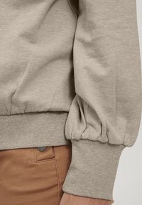 Svetlo bež pulover z teksturirano površino, ki ima napihnjene rokave in prilegajoč rob, v kombinaciji z rjavimi hlačami z gumbom.