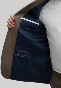 Braunes Wollanzugjackett mit einem satinschwarzen Futter. Es verfügt über eine weiße Trimmpocket und Etiketten, die den Stofftyp und die Herkunft anzeigen.