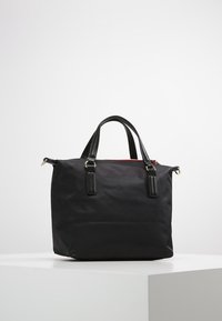 Sort rektangulær håndtaske med to korte læderhåndtag og guldfarvet hardware, stående på en hvid sokkel foran en ensfarvet baggrund.