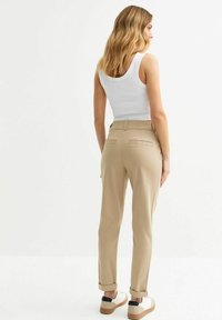 Beige Hosen mit geradem Schnitt und umgeschlagenen Säumen, ausgestattet mit zwei Gesäßtaschen. Kombiniert mit einem weißen Tanktop und zweifarbigen Sneakern.