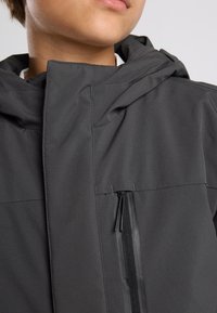 Musta huppari, jossa on sileä pinta, vasemmassa rintataskussa vetoketju ja minimalistiset suunnitteluominaisuudet.