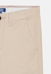 Pantalons en coton beige avec une texture lisse, dotés de poches avant et d'un bouton noir à la taille. Surpiqûres contrastées le long de la ceinture.