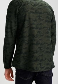 Długi rękaw, ciemnozielona koszula z wzorem kamuflażu, z zaokrąglonym dołem i mankietami na guziki, wykonana z miękkiej, teksturowanej tkaniny.