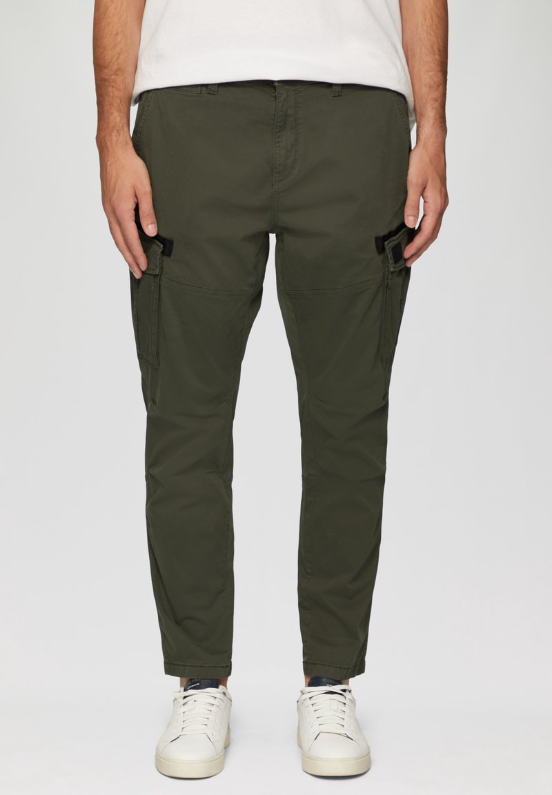 Homme portant un pantalon cargo vert olive avec des poches sur les côtés et des baskets blanches, debout devant un fond uni.