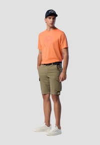 T-shirt arancione con logo, pantaloni cargo beige, sneakers bianche e berretto blu con testo ricamato. Outfit casual adatto per il clima caldo.