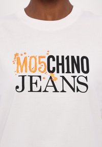 Bílý bavlněný tričko s výrazným černým textem "CH1NO JEANS" a oranžovým "M05" se skvrnitým efektem, zaměřující se na detaily grafického designu.