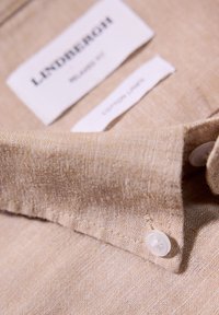 Colletto di camicia in lino beige con un pulsante visibile, dotato di etichetta del marchio "Lindbergh" e etichetta di cura, tessuto testurizzato con una trama sottile.