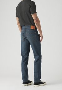 Homme portant un jean Levi's bleu foncé, un t-shirt noir et des baskets noires, debout de dos, montrant des tatouages aux coudes.