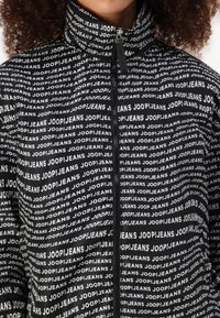 Juodas švarkas su pakartotiniu baltu tekstu "JOOP!JEANS." Aukšta apykaklė, užtrauktukas priekyje ir lygi tekstūra.