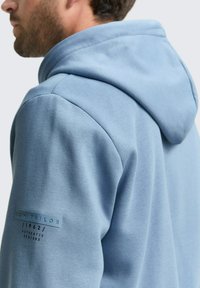 Lichtblauwe hoodie van zacht materiaal. Heeft een gestructureerde capuchon en een label op de linker mouw. Glad textuur met minimale details.