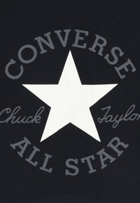 Μαύρο ύφασμα με ένα μεγάλο λευκό αστέρι στο κέντρο, με τις λέξεις «CONVERSE» και «ALL STAR» σε γκρι, καθώς και το «Chuck Taylor» σε γραμματοσειρά χειρόγραφου στυλ.