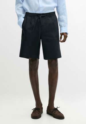 Uomo che indossa pantaloncini neri con coulisse, camicia a maniche lunghe azzurro chiaro e mocassini marroni, in piedi davanti a uno sfondo bianco semplice.