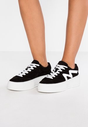 Nogi w czarnych zamszowych platformowych trampeczkach z białym logo w kształcie zygzaka i grubymi białymi gumowymi podeszwami, stojące na białej powierzchni.