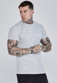 Grijs textuur kortemouwen t-shirt met een aansluitend ontwerp en een ronde halslijn, gedragen met donkere spijkerbroeken. Zichtbare tatoeages op de armen.