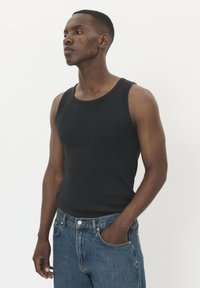 Uomo in canotta nera senza maniche e jeans blu, in piedi con una mano in tasca, che guarda leggermente a sinistra su uno sfondo bianco.