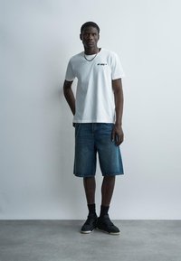 Mustanahaline mees seisab tavalise seina ees, seljas valge Off-White T-särk, põlvpikad teksapüksid, mustad sokid, mustad tossud ja ketiga kaelakee.