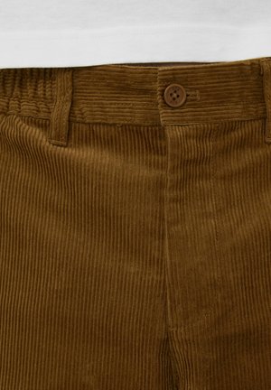 Pantalon en velours côtelé marron avec texture à côtes verticales, doté d'une fermeture à boutons et de passants pour ceinture. Le matériau semble doux et résistant.