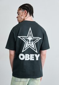 Black cotton t-shirt featuring a large white star graphic with a face, and the word "OBEY" in bold white letters on the back.