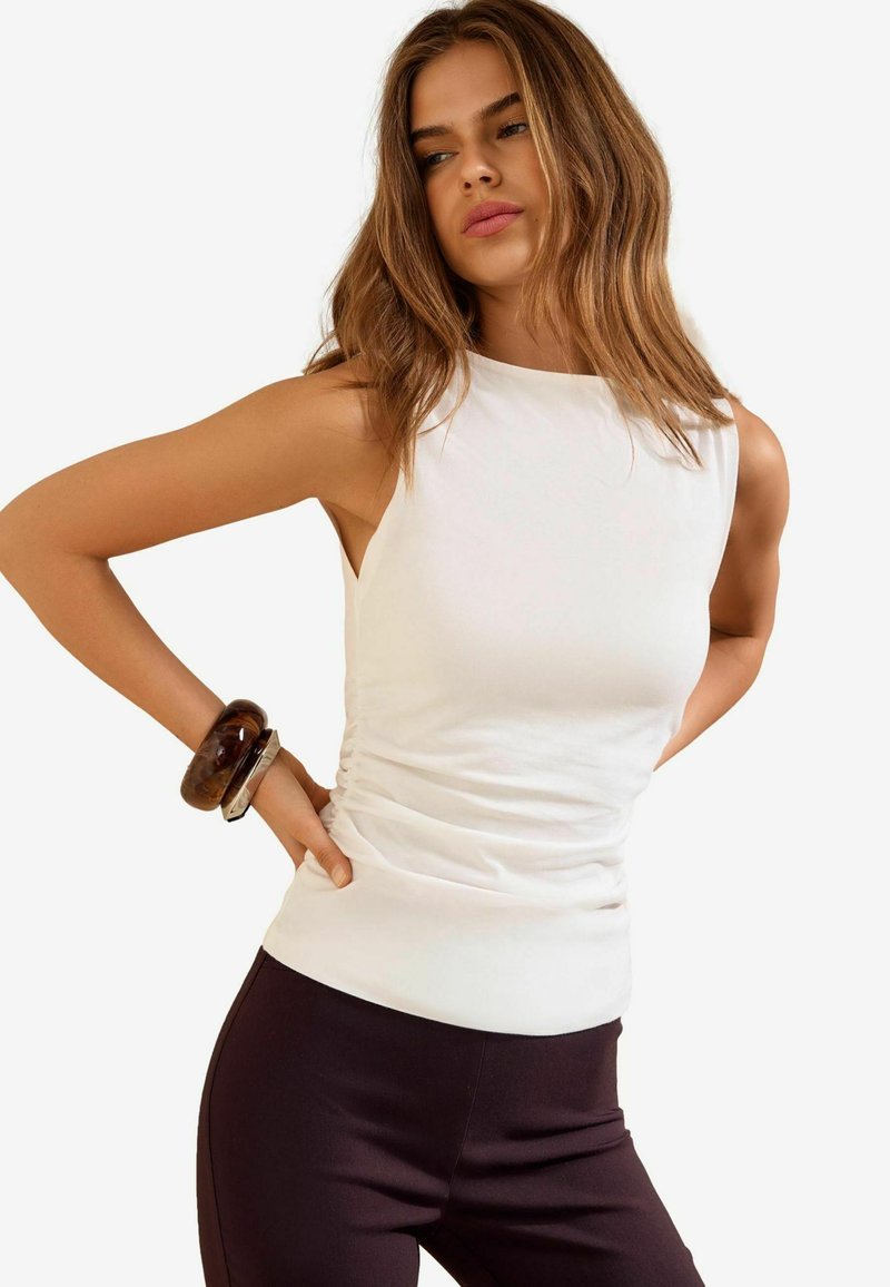 Woman with wavy brown hair wearing a sleeveless white top and dark pants, posing with hands on hips, wearing chunky bracelets on left wrist.