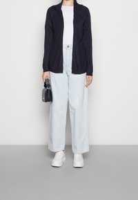 Tmavě námořnický žebrovaný kardigan s šálovým límcem, doplněný světle modrými džínami s širokými nohavicemi a bílými teniskami. V jedné ruce drží malou černou tašku.