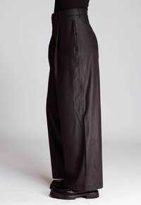 Side view of a person wearing high-waisted, wide-leg black leather pants and chunky black shoes on a plain background.