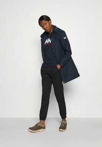 Tamsiai mėlynas vėjuotas švarkelis su gobtuvu, ilgomis rankovėmis ir logotipo detale, derinamas su tamsia marškinėliais, juodomis siaurėjančiomis kelnėmis ir ruda sportine avalyne.