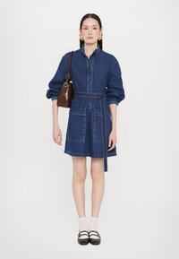 Rochie din denim cu mâneci lungi tip puff, închidere cu nasturi în față, două buzunare frontale, talie cu curea și o geantă marron închis. Șosete albe și pantofi negri.