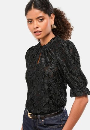 Woman with curly hair wearing gold earrings, a black floral lace blouse with puff sleeves, dark jeans, and a black belt with gold buckle.
