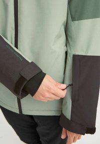 Wasserdichte Jacke mit einem hellgrünen Oberteil und schwarzen unteren Ärmeln, verstellbaren Bündchen, einer Reißverschlusstasche und strukturierten Stoffbereichen.