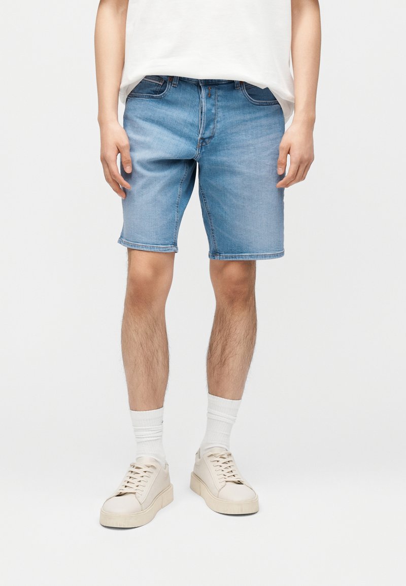 Άνδρας που φοράει ανοιχτόμπλε τζιν σορτς, λευκές κάλτσες και εκρού αθλητικά παπούτσια, με φόντο ένα απλό λευκό τοίχο.