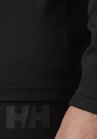 Zwarte thermoshirt van een textuur- en ademend materiaal. Heeft een zichtbaar logo "HH" in matte afwerking, wat het sportieve ontwerp benadrukt.