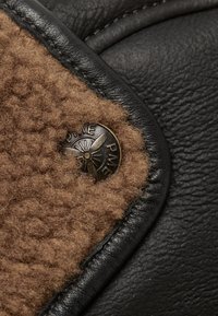 El borde de cuero negro se encuentra con un suave y esponjoso shearling marrón claro, adornado con un botón circular de bronce que presenta un diseño de hélice.