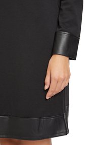 Main de femme reposant à côté d'une robe noire avec des bordures brillantes en simili-cuir sur le poignet et l'ourlet, sur fond blanc.