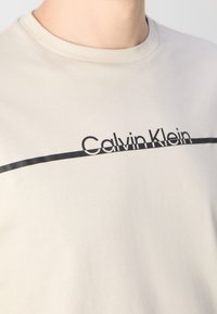 Gros plan sur une personne portant un t-shirt beige à col rond avec le texte « Calvin Klein » et une ligne noire horizontale traversant la poitrine.