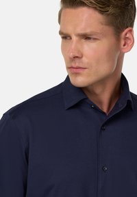 Muž so svetlou pokožkou a krátkymi hnedými vlasmi, oblečený v tmavomodrej košeli na gombíky, pozerajúci sa mierne doľava s neutrálnym výrazom.