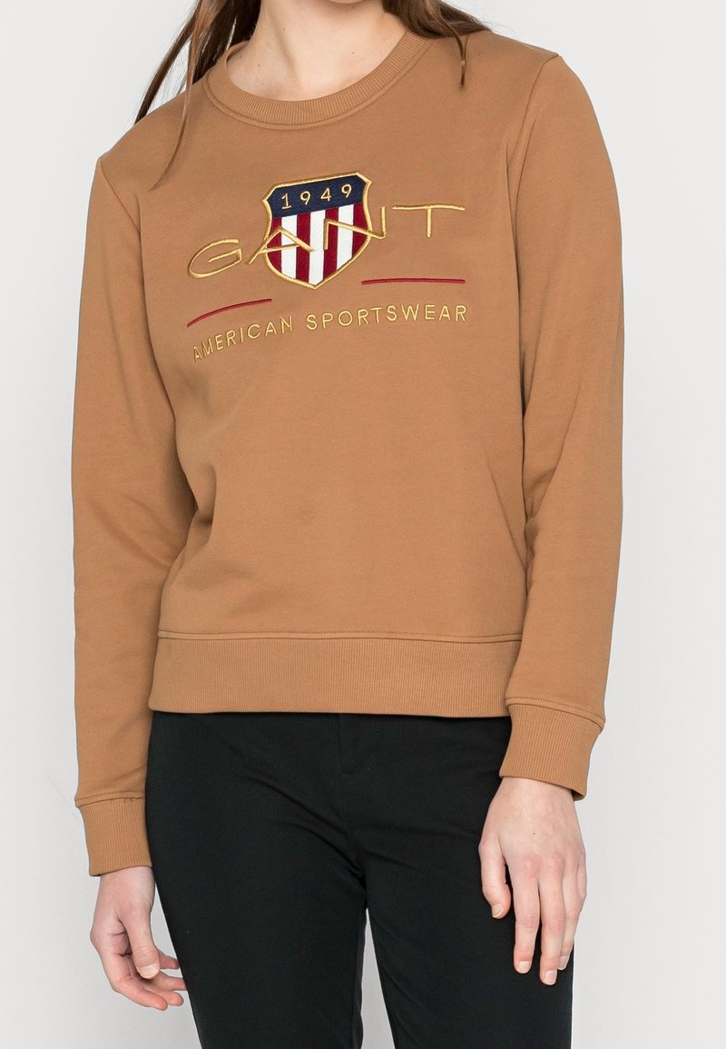 Brauner Sweatshirt mit Rundhalsausschnitt, ausgestattet mit einem gestickten Wappenlogo in Rot, Blau und Gold, kombiniert mit schwarzen Hosen.
