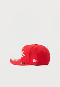 Casquette de baseball rouge avec une visière plate, broderie blanche sur le devant et un petit logo sur le côté. Fabriquée en tissu avec une texture lisse.