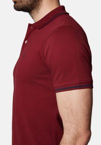 Burgundowa koszulka polo o teksturowanym wykończeniu, z dwiema białymi guzikami oraz kontrastującymi niebieskimi i czerwonymi paskami na kołnierzyku i rękawach.