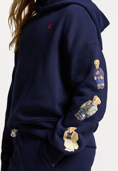 Námořnická modrá mikina s kapucí vyrobená z bavlněné směsi, na pravém rukávu s třemi ilustracemi medvědů v různých oblecích a detailech.