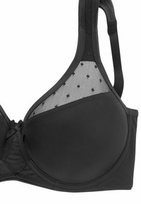 Zwarte bh met voorgevormde cups, een mesh paneel met stippen, verstelbare bandjes en een strikdetail in het midden. Gladde textuur met een gestructureerd ontwerp.