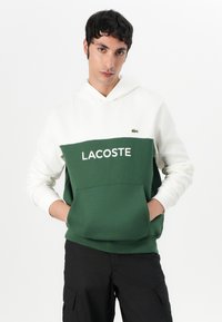 Roheline ja valge kapuutsiga sviitriga, millel on ees tasku ja "LACOSTE" logo. Pehme kangas, kontrastsete värvidega ja mugav lõige.