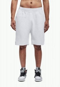 Mann in hellgrauen knielangen Shorts und weißen High-Top-Sneakern mit schwarzen Schnürsenkeln vor einem weißen Hintergrund.