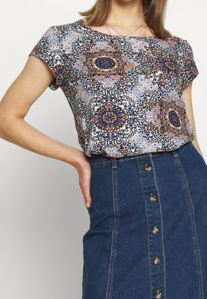 Blusa a maniche corte caratterizzata da un motivo floreale e geometrico multicolore su uno sfondo chiaro, abbinata a una gonna di jeans blu con bottoni.