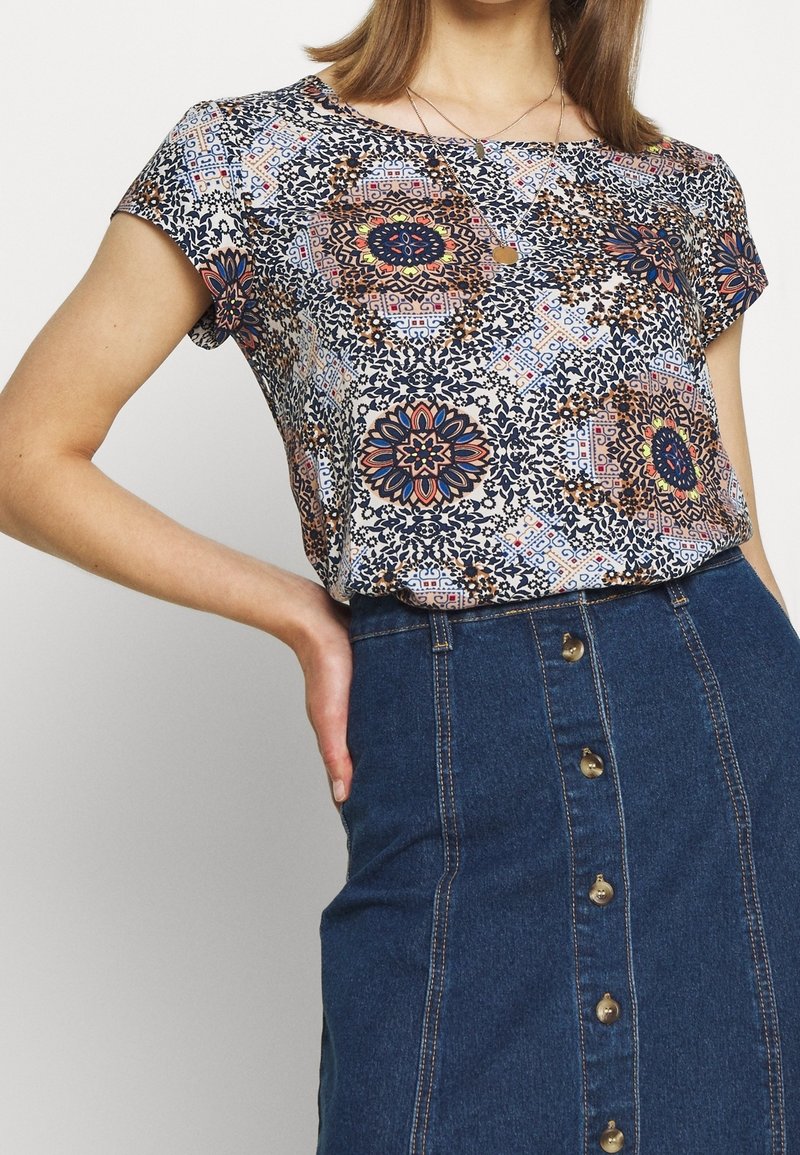 Blusa de manga corta con un patrón floral y geométrico multicolor sobre un fondo claro, combinada con una falda de mezclilla azul con botones.