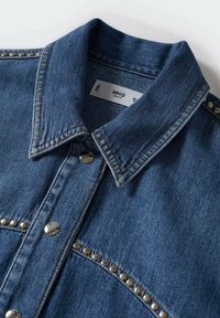 Col et haut de la veste en jean bleu avec boutons-pression argentés et détails de clous en métal le long des coutures.