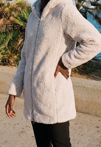 Personne portant une veste à capuche beige clair en tissu pelucheux avec fermeture éclair, debout à l'extérieur près de plantes de palmier et d'une barrière en béton.