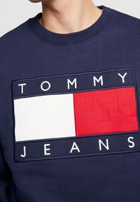 Sudadera azul marino con un logo rectangular que presenta bloques en rojo, blanco y azul. "TOMMY" y "JEANS" están bordados en letras blancas.