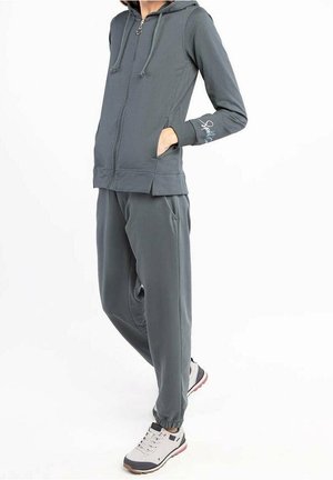 Persona con sudadera con capucha gris oscuro con cremallera y bolsillos, y pantalones jogger a juego, combinados con zapatillas deportivas gris claro y blancas.