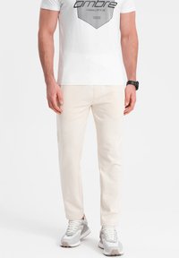 Beige Baumwollhose mit geradem Schnitt und seitlichen Taschen, kombiniert mit einem weißen Grafik-T-Shirt und grauen Sneakern. Schlichtes Design.